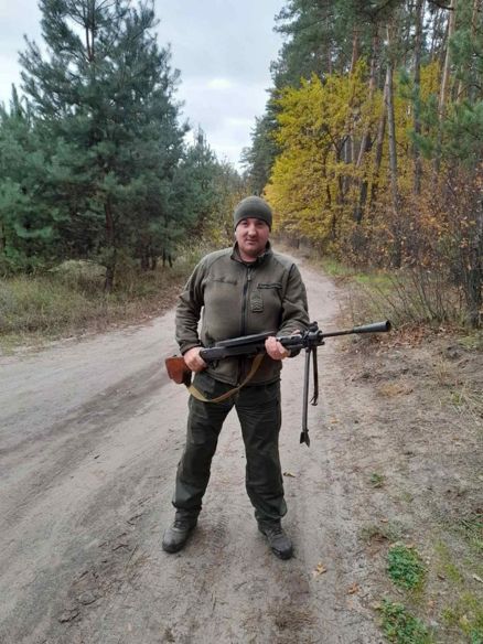 Чоловік у камуфляжі тримає автомат на ґрунтовій дорозі в лісі.