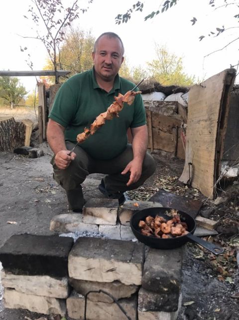 Чоловік у зеленій сорочці тримає шашлик на шпигу біля вогнища з казана.