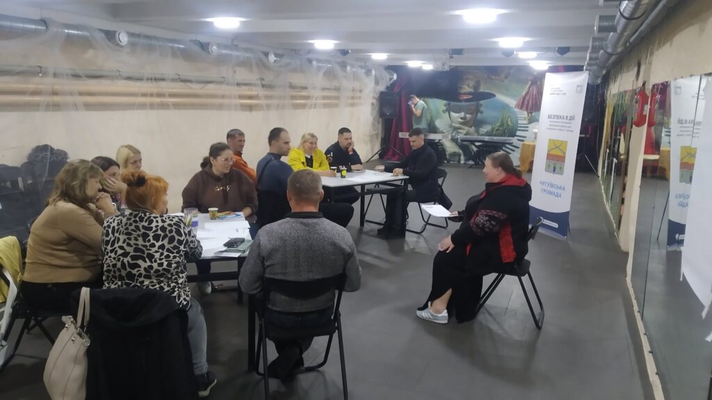 Група людей сидить за довгим столом під час зустрічі, поруч стоїть банер з написом «ВІДКРИВАЄМО В ДІЙ ЧУТЛИВУ ГРОМАДУ».