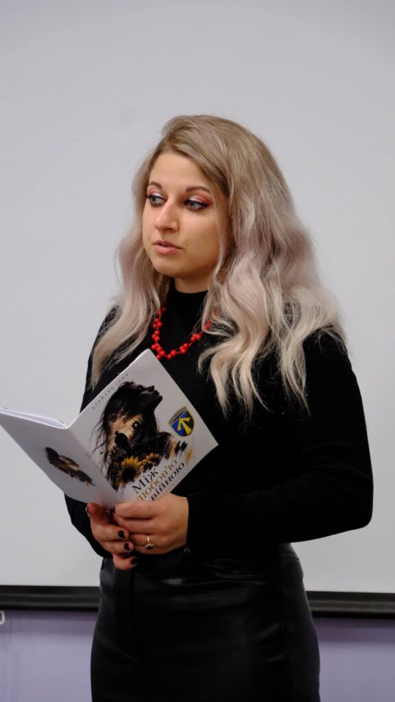 Жінка з сивим волоссям у чорному одязі тримає відкриту книгу з написом «Міфологія» та зображенням птаха.