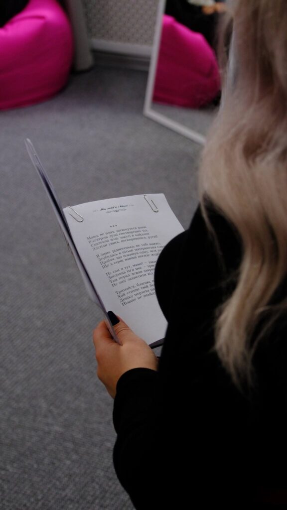 Жінка з blond волоссям у чорному одязі читає аркуш паперу з текстом, тримаючи його в руці.