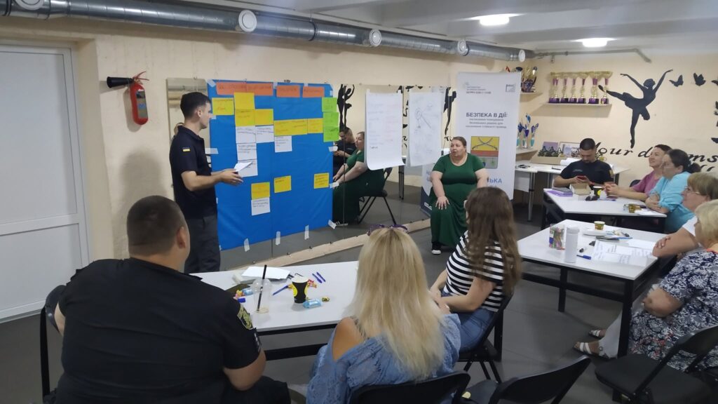 Чоловік у формі проводить тренінг для групи людей, які сидять за столами, з дошками з липкими нотатками та банером «Безпека в дії» на тлі.