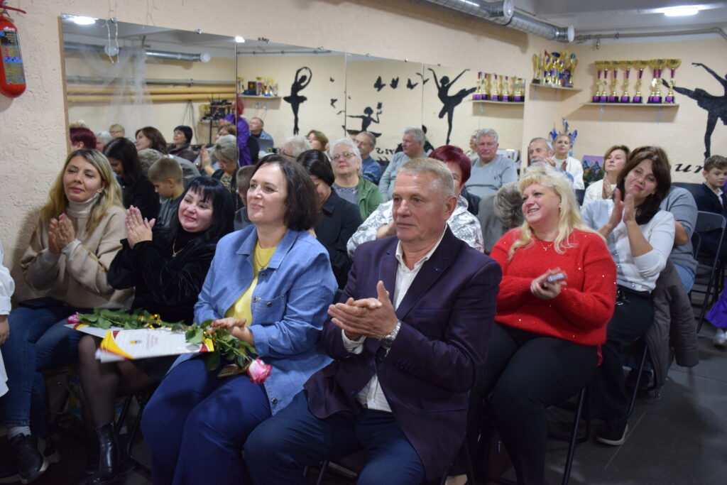 Публіка в залі з дзеркалами та кубками плескає оплесками, а на передньому плані жінка тримає букет квітів.