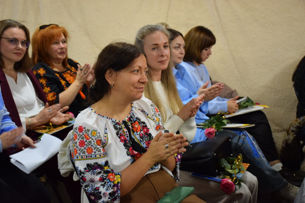 Група жінок, одягнених у традиційні вишиті сорочки, сидить у залі та оплескує.