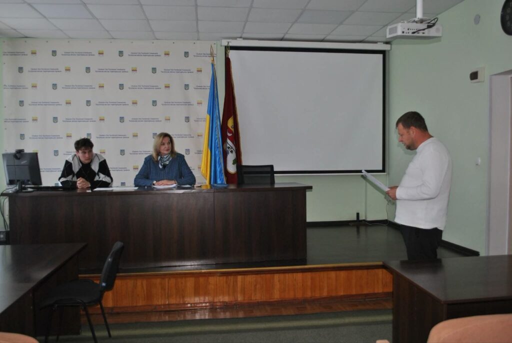 Чоловік у білій сорочці читає документи, стоячи біля столу, де сидять дві жінки, на тлі прапорів та екрана.