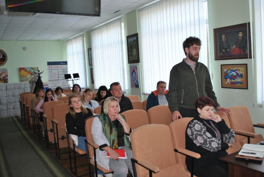 Чоловік стоїть біля ряду аудиторних крісел, де сидять слухачі, що уважно слухають лекцію або презентацію.