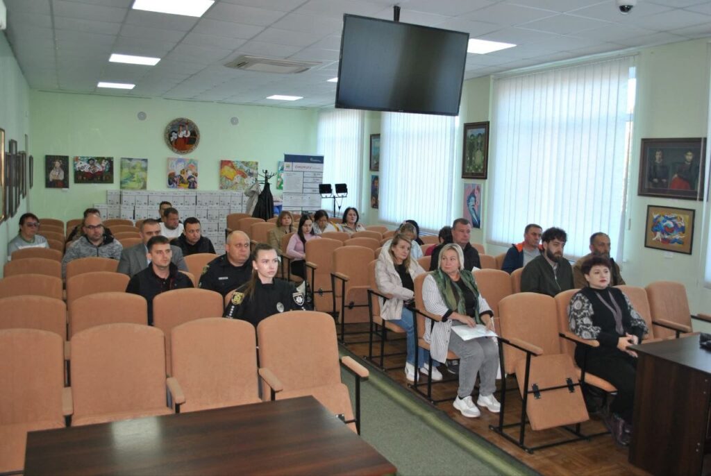 Група людей сидить у залі з рядами крісел, слухаючи виступ біля трибуни.