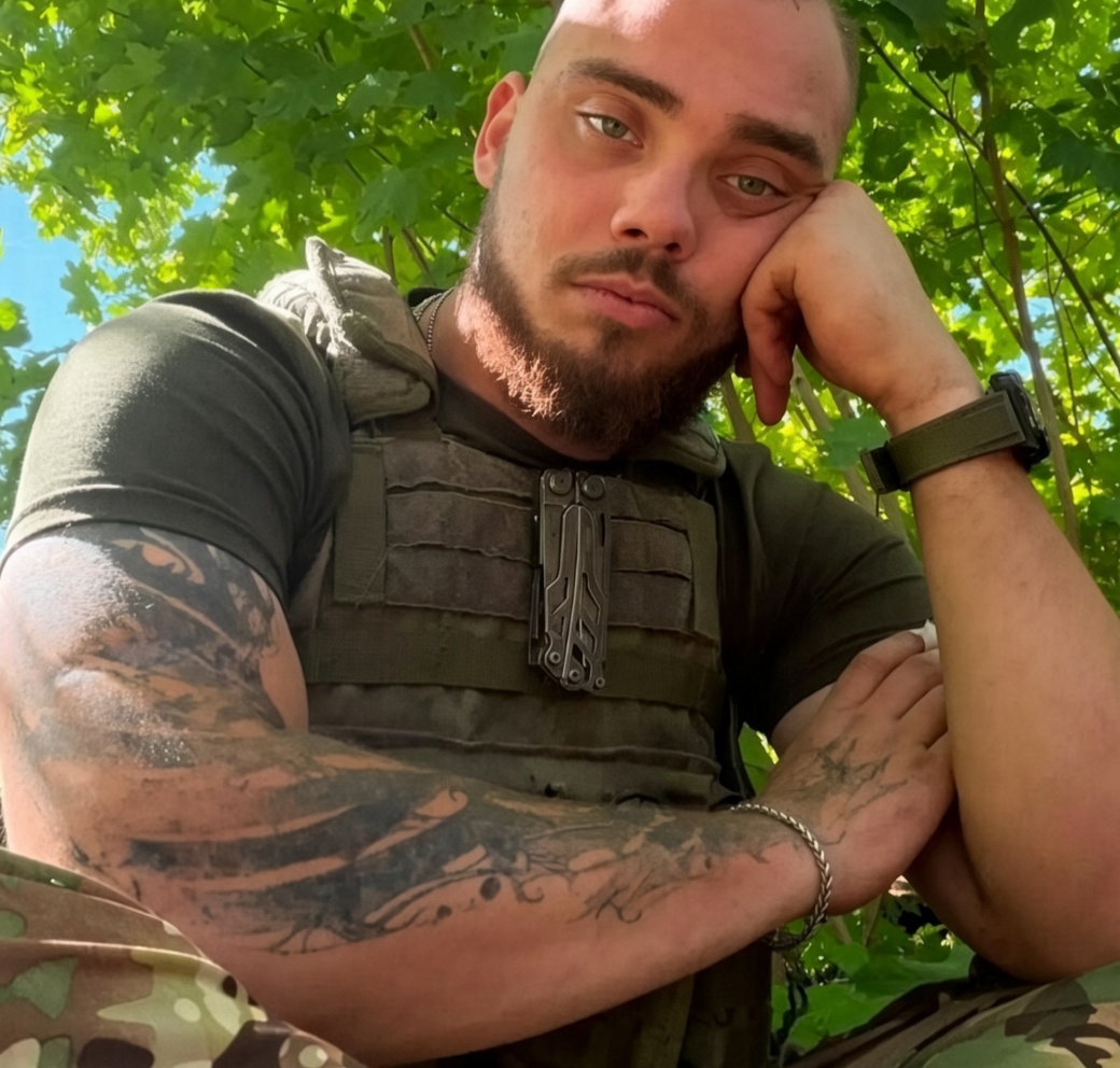 Чоловік у тактичному жилеті та камуфляжі сидить під деревами, спираючись рукою на щоку.
