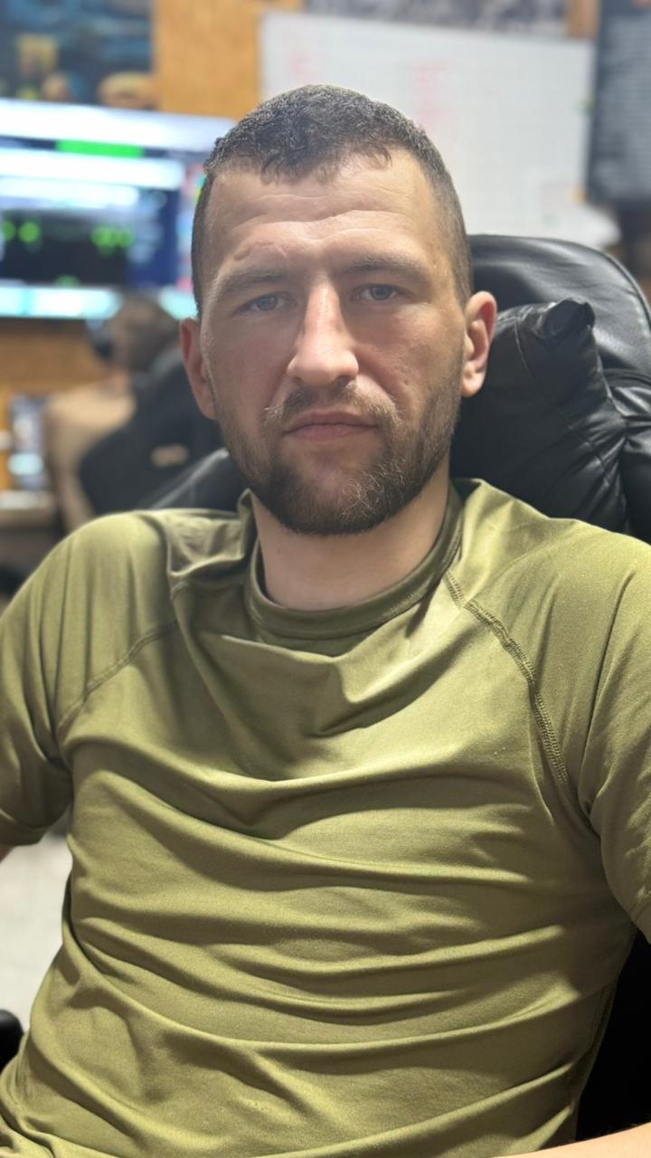 Чоловік із коротким волоссям та бородою в оливковій футболці сидить у чорному кріслі в офісі.